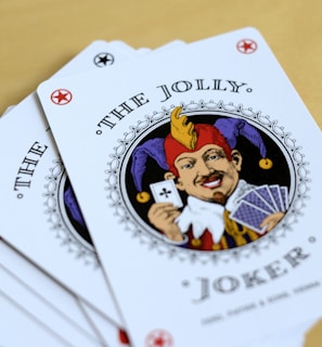 A fan of playing cards spread out on a light wooden table, featuring prominently is a Joker card with intricate illustrations. The Joker has a colorful jester hat and is holding a card and a hand of other cards.