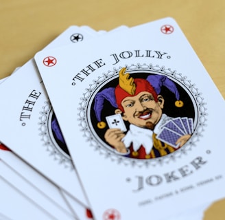 A fan of playing cards spread out on a light wooden table, featuring prominently is a Joker card with intricate illustrations. The Joker has a colorful jester hat and is holding a card and a hand of other cards.