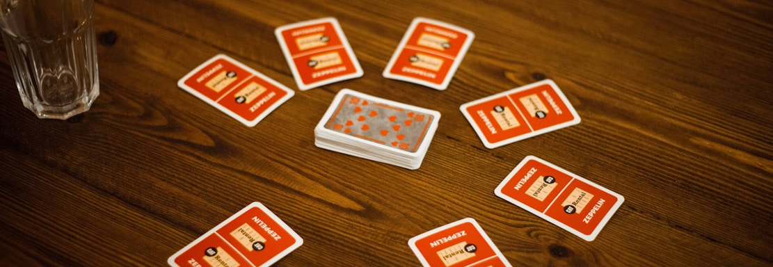 A wooden table with several playing cards arranged in a circle around a stack of cards in the center. Each card has a red back with text and illustrations. Two drinking glasses, one with a clear liquid, are also on the table, with a person's arm visible in the background.
