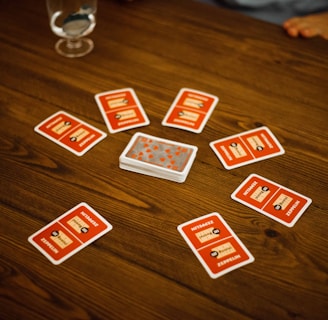 A wooden table with several playing cards arranged in a circle around a stack of cards in the center. Each card has a red back with text and illustrations. Two drinking glasses, one with a clear liquid, are also on the table, with a person's arm visible in the background.