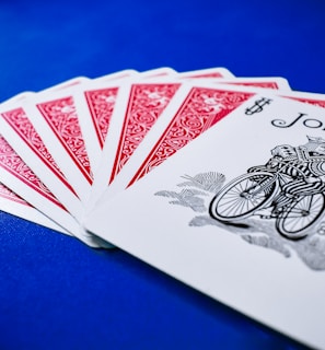 A set of playing cards spread out on a vibrant blue surface, highlighting a joker at the forefront. The backs of the other cards feature a red intricate design with white borders.