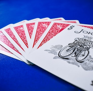 A set of playing cards spread out on a vibrant blue surface, highlighting a joker at the forefront. The backs of the other cards feature a red intricate design with white borders.