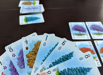 A hand holds several playing cards with tree illustrations and different numbers, set against a dark table. Additional cards are arranged in rows on the table, showing various tree images and numbers.