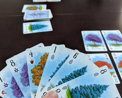 A hand holds several playing cards with tree illustrations and different numbers, set against a dark table. Additional cards are arranged in rows on the table, showing various tree images and numbers.