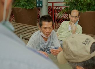 Several individuals gather outdoors, sitting on benches and engaging in a game of cards. The focal point is on a man holding a fan of playing cards, with others observing or participating. Potted plants and a red railing are visible in the background, contributing to the setting's casual and social atmosphere.