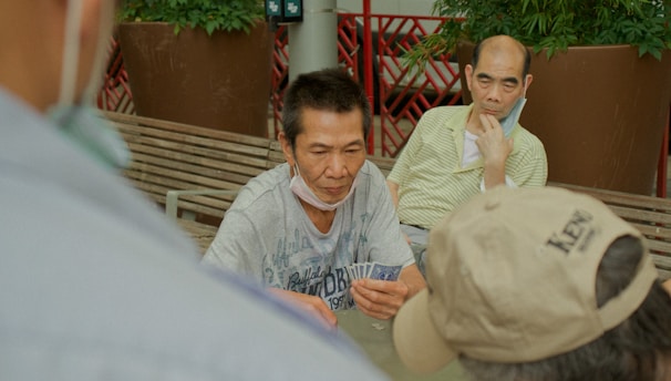 Several individuals gather outdoors, sitting on benches and engaging in a game of cards. The focal point is on a man holding a fan of playing cards, with others observing or participating. Potted plants and a red railing are visible in the background, contributing to the setting's casual and social atmosphere.