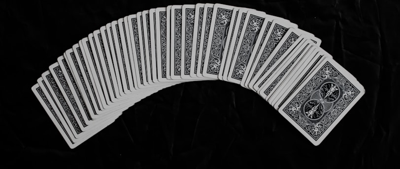 A neatly arranged spread of playing cards with an intricate design on their backs, set against a dark fabric background. The cards form a gentle curve, creating a sense of symmetry and order.