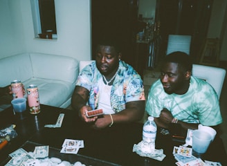 Two individuals are sitting at a table engaged in a card game. Various playing cards, poker chips, and items are scattered on the table. Arizona tea cans and drinks are present, and both people appear relaxed and focused.