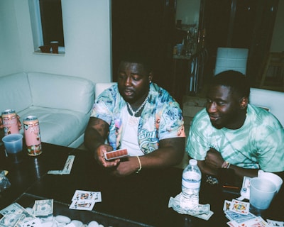 Two individuals are sitting at a table engaged in a card game. Various playing cards, poker chips, and items are scattered on the table. Arizona tea cans and drinks are present, and both people appear relaxed and focused.