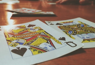 Playing cards are spread out on a wooden table, with the Queen and King of Spades prominently visible. The focus is on the vibrant colors and detailed designs of the cards, while other cards and hands are slightly blurred in the background, suggesting a game in progress.
