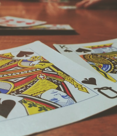 Playing cards are spread out on a wooden table, with the Queen and King of Spades prominently visible. The focus is on the vibrant colors and detailed designs of the cards, while other cards and hands are slightly blurred in the background, suggesting a game in progress.