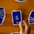 A hand holds a playing card with Arabic text printed on it. Several other cards are spread out on a wooden surface in different positions.