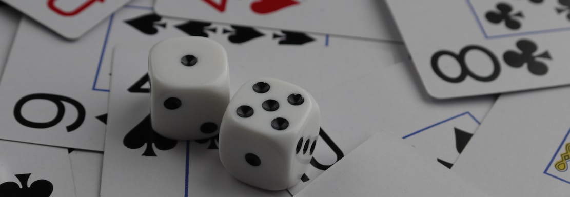 A collection of playing cards spread out, featuring various suits such as hearts, clubs, and spades. Two white dice with black dots are placed prominently on top of the cards.
