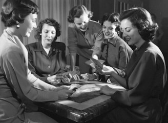 Five women are gathered around a table, engaged in a card game. Some are looking at their cards intently, while others are smiling and interacting with each other. The setting suggests a social and relaxed atmosphere.