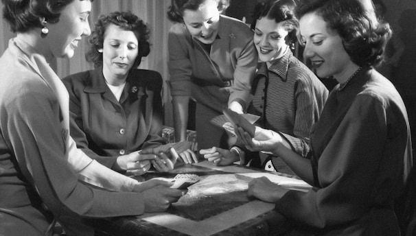 Five women are gathered around a table, engaged in a card game. Some are looking at their cards intently, while others are smiling and interacting with each other. The setting suggests a social and relaxed atmosphere.