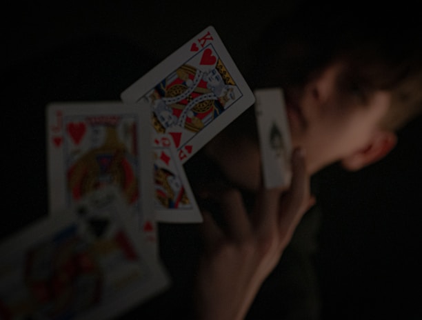 A person holding a playing card in their hand, with other playing cards floating in mid-air. The background is dark, highlighting the contrast between the cards and the surroundings.